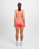 Back view of woman wearing coral Nexletica Impulse High Neck Sports Bra and Impulse High Waist Shorts 5 inch showing waistband detail and logo